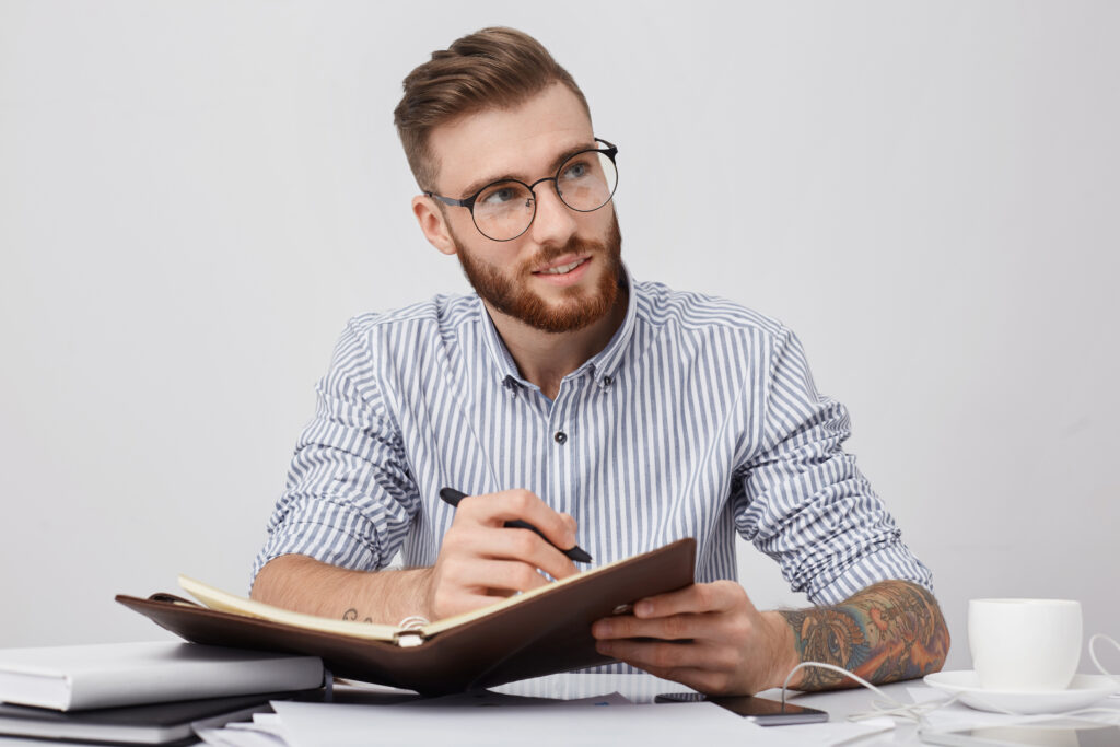 thoughtful male manager in round spectacles, wears formal shirt, writes in notebook as sits at work place, generates new ideas for creative future project. people, business, occupation concept
